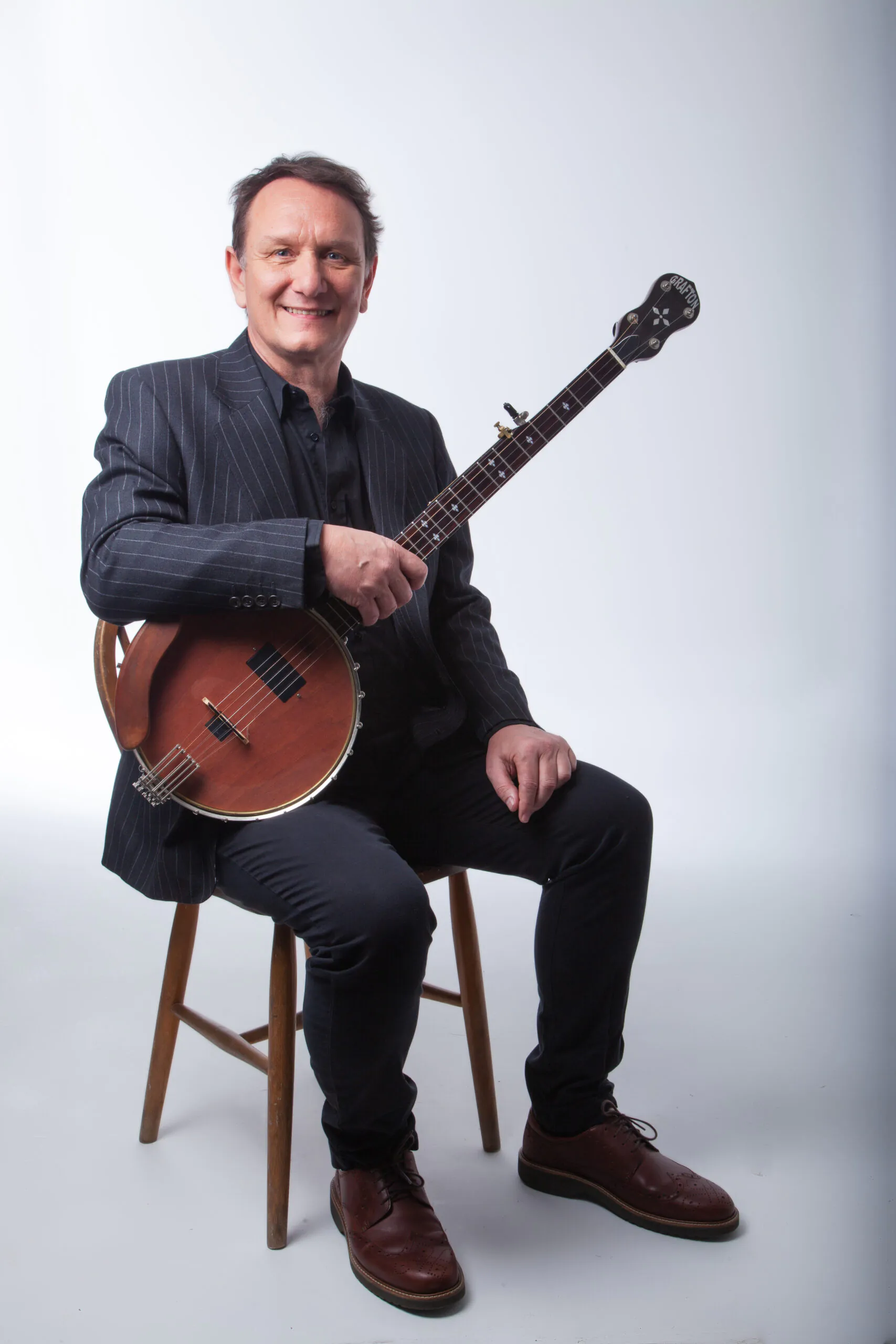Joff Lowson in a pin stripe suit with his banjo on his knee smiling at the camera